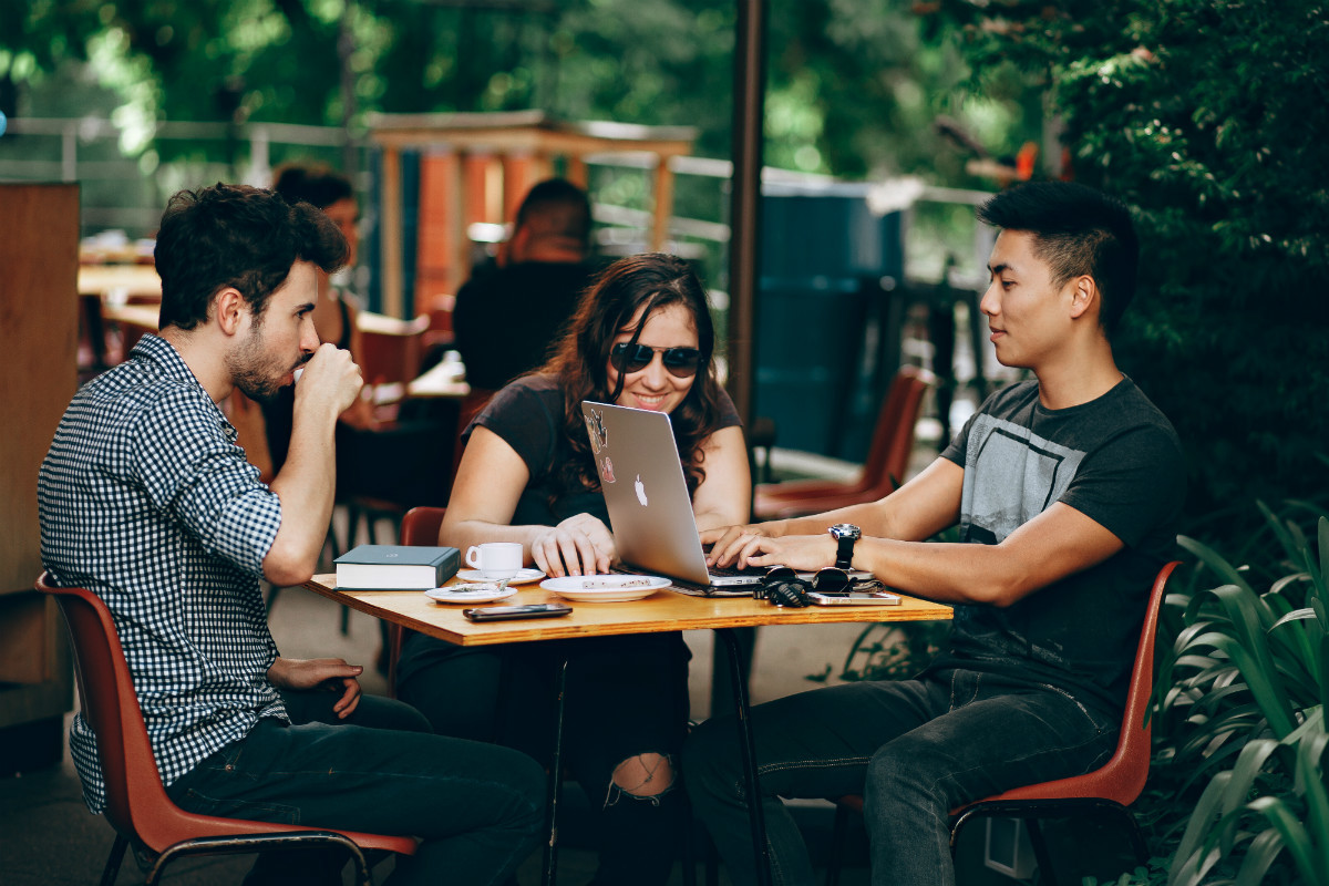 Three young people with laptops at a cafe discuss their first job after coding bootcamp Three young people with laptops at a cafe discuss their first job after coding bootcamp